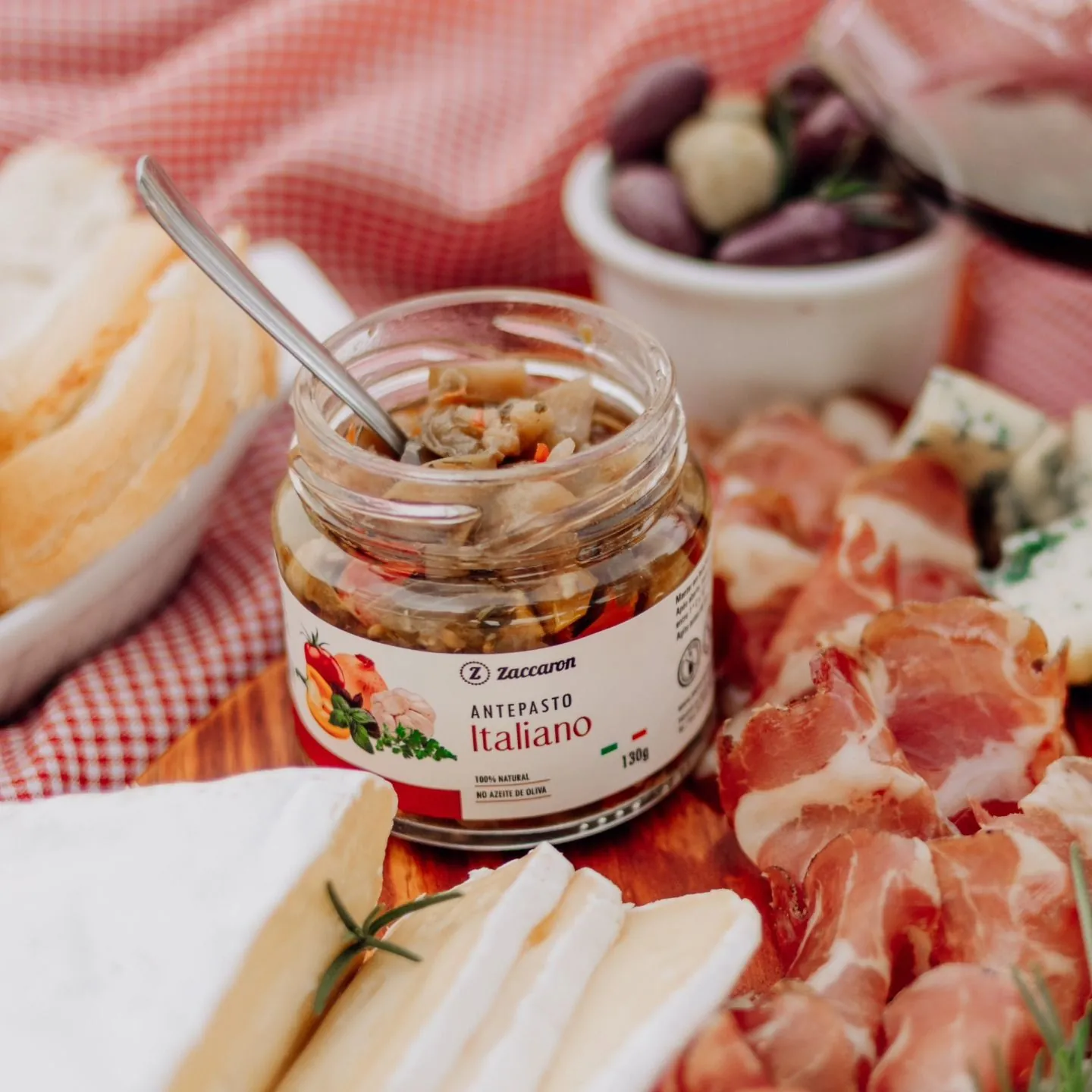 Pote de Antepasto Italiano Zaccaron aberto sobre toalha xadrez vermelha, com queijos e frios