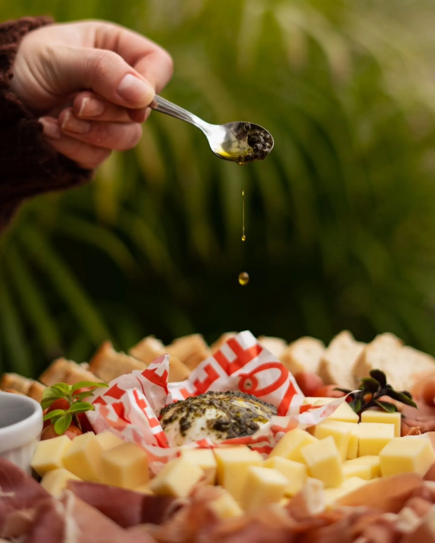 Mão derramando azeite com uma colher sobre tábua de queijos, pães e frios
